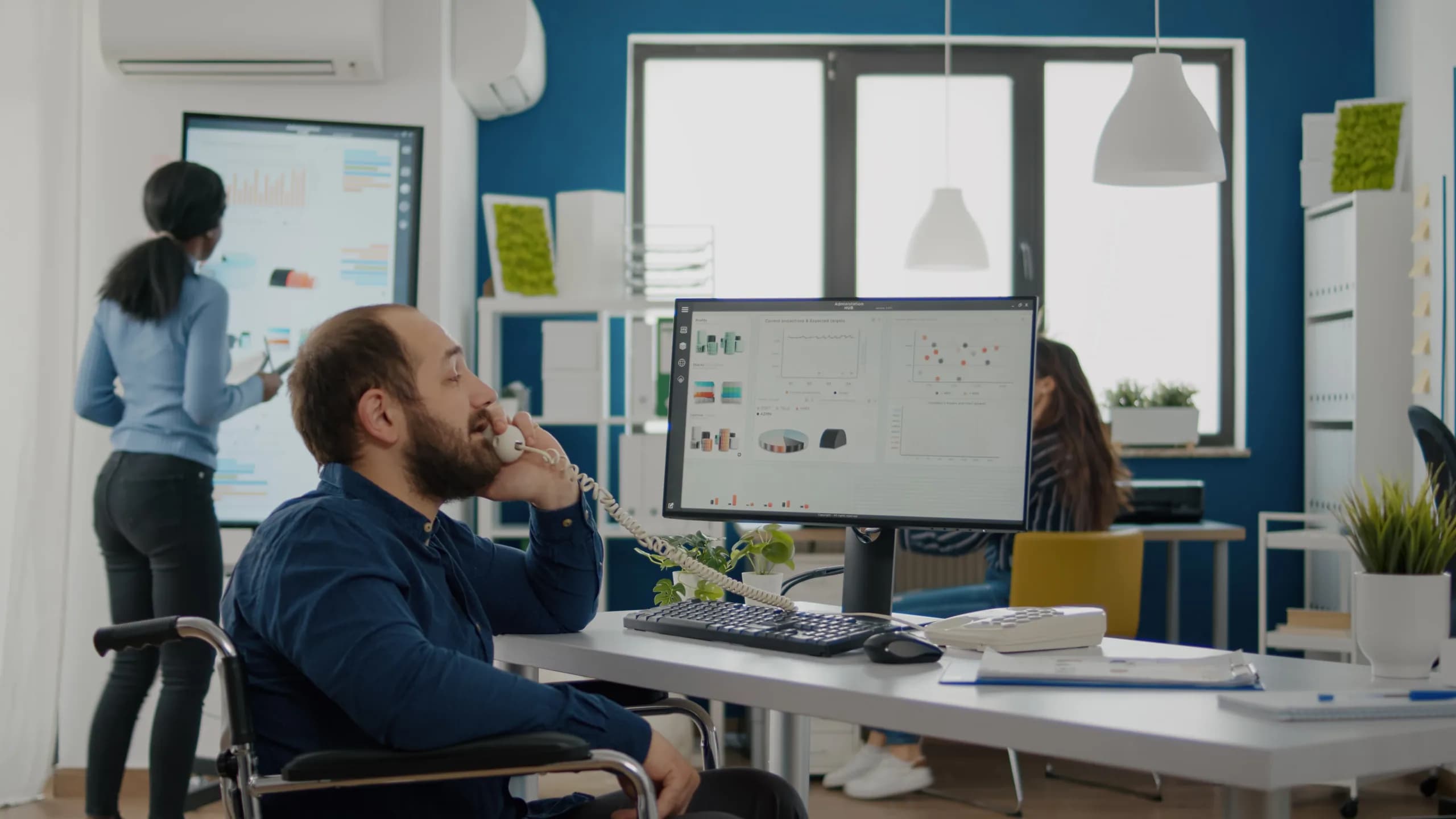 An operations manager on a desk phone reviewing an administration dashboard of stock and order charts, with colleagues working behind him